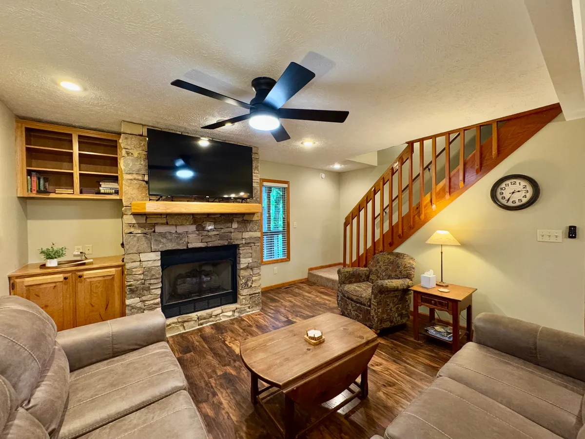 This image features a spacious living room with a prominent stone fireplace, cozy seating arrangements, a mounted TV, a wooden staircase, and a central coffee table. The ambiance is warm and welcoming, perfect for group gatherings, game nights, or relaxing by the fire.