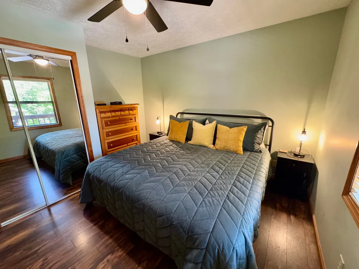 A spacious bedroom featuring a king-sized bed dressed in a blue quilt with yellow pillows, a wooden dresser, and a mirror reflecting the tranquil ambiance of the room. The sunlight filters softly through the window, creating an inviting and restful setting.