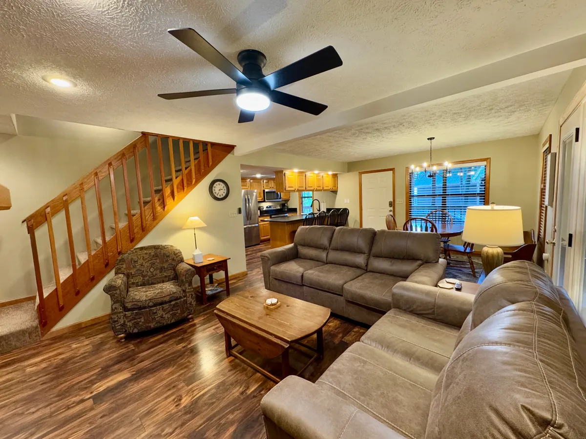 A spacious, inviting living room featuring a large sectional sofa, a wooden coffee table perfect for board games or snacks, a decorative lamp, and a visible staircase leading upstairs. The warm, homely decor encourages socializing and group activities, with plenty of room for multiple generations to gather comfortably.