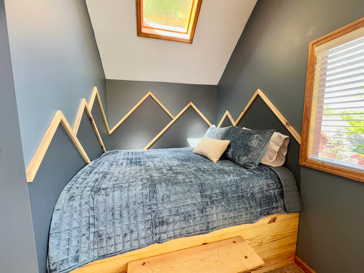 A serene bedroom featuring plush bedding, unique geometric wood accents, and a skylight overhead. The gentle daylight creates a calm and dreamy atmosphere, suggesting restful nights and opportunities to watch the stars. The inviting space encourages couples to curl up and enjoy tranquil moments together.