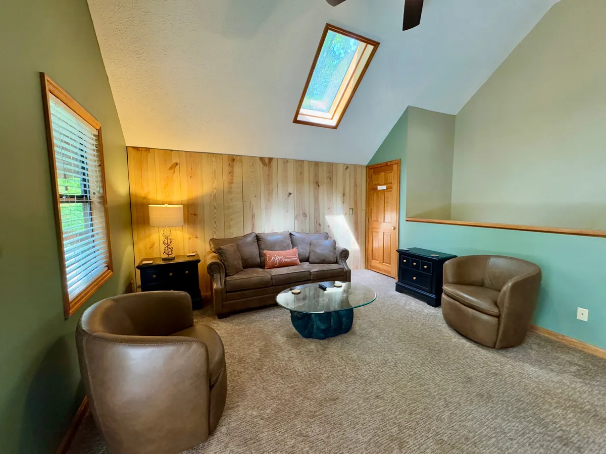 A cozy loft area featuring a sofa with cushions, two armchairs positioned for conversation or relaxation, a glass coffee table, and an overhead skylight that lets in abundant natural light. The space radiates warmth and flexibility, ideal for reading, socializing, or lounging.