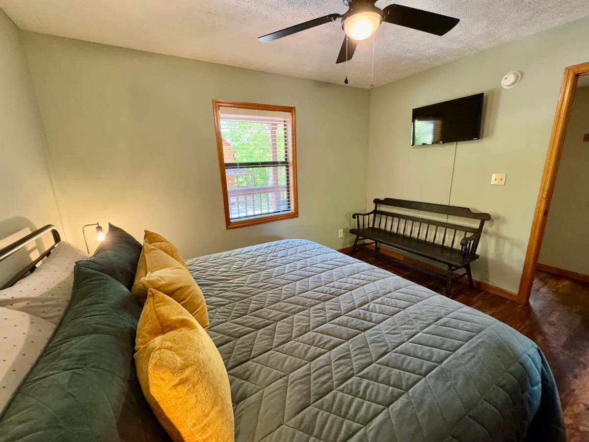 A comfortable bedroom featuring a plush bed with soft pillows, a window allowing natural light to fill the space, a wooden bench, and a mounted TV for entertainment. The room exudes a cozy, comfortable vibe, ideal for relaxing or enjoying quiet moments.