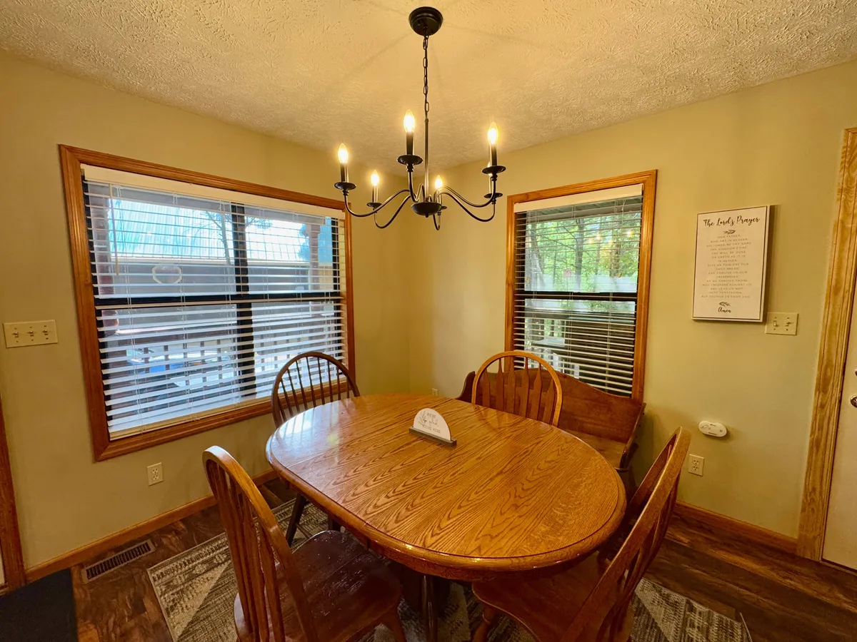 A large wooden dining table is set beneath a chandelier and surrounded by windows, creating a bright, inviting setting for group meals, family traditions, and lively conversations.